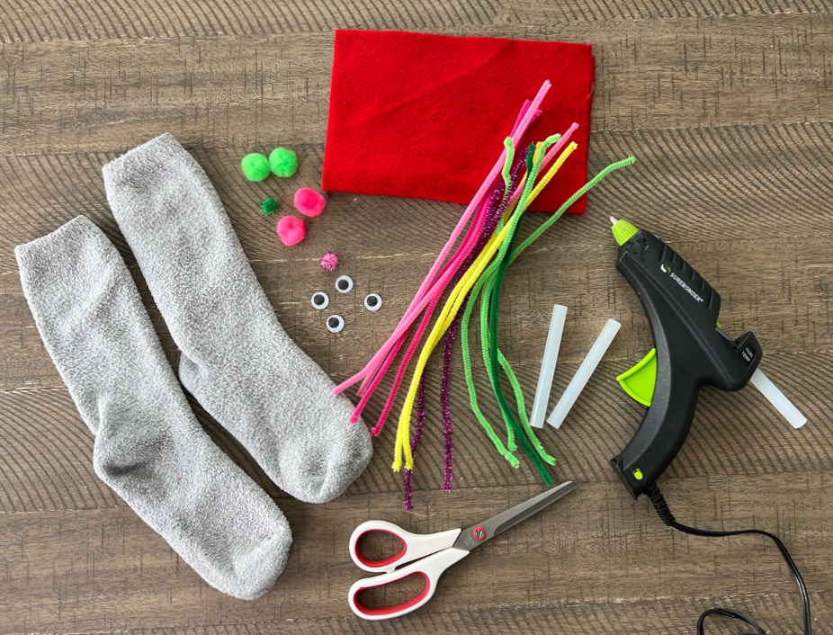 craft supplies including a sock, felt material, googly eyes, craft pipe cleaners, pom poms, scissors, hot glue gun.